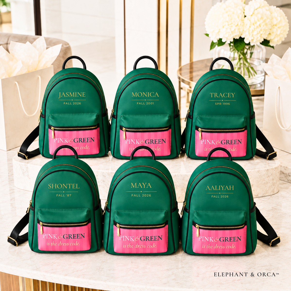 Six personalized green mini backpacks arranged on a marble surface, each with a bright pink front pocket and gold embroidered names and text. The scene includes gift bags and white flowers in the background, making it a styled product display.